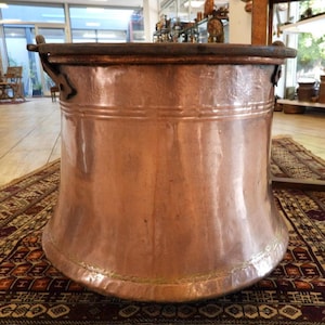 May include: A large, cylindrical copper vessel with a wide, flared base and a dark, reinforced rim. The exterior surface features a hammered texture with visible horizontal bands and metal side handles attached with decorative rivets.