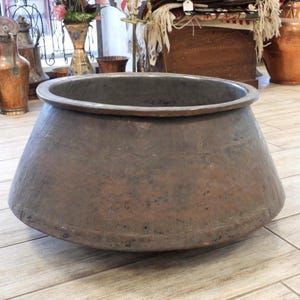 May include: A large, wide-mouthed copper vessel with a tapered, conical body and a smooth, rounded rim. The exterior surface features a weathered, dark brown patina with visible signs of age, including subtle surface texture and metallic oxidation.