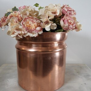 May include: A large, copper-coloured vase filled with faux flowers. The flowers are a mix of pink and cream-coloured blooms. The vase has a cylindrical shape with a slightly wider top and bottom.
