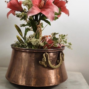 Könnte beinhalten: Ein gehämmerter Kupfertopf mit breitem Boden und zwei Messinggriffen, gefüllt mit einem Arrangement aus rosa Lilien, grünem Blattwerk und kleinen weißen Blüten. Die Metalloberfläche weist durchgehend eine texturierte, genoppte Optik auf.