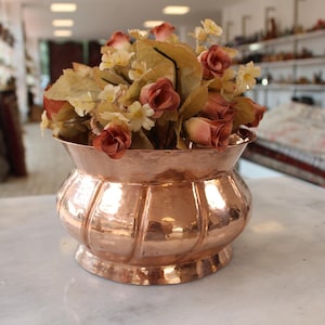 May include: A copper vase with a hammered finish, featuring a fluted body and a flared rim. It contains a bouquet of artificial flowers, including peach roses and small yellow flowers. The vase is on a white marble surface.