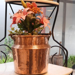 May include: A hammered copper bucket with two side handles, filled with an arrangement of artificial pink lily flowers, green foliage, and small orange berries. The bucket has a wide, cylindrical body with a slightly tapered neck and a metallic finish.