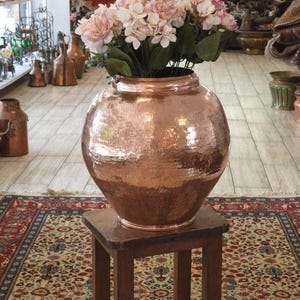 Op de afbeelding: Een grote, ronde vaas van gehamerd koper met een brede buik en een smalle hals, gevuld met een boeket lichtroze en witte kunstbloemen. De vaas heeft een gepolijst, reflecterend metalen oppervlak met zichtbare handgehamerde inkepingen.