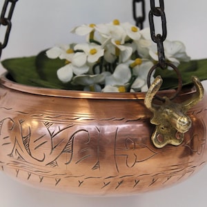 Könnte beinhalten: Ein runder hängender Pflanztopf aus Kupfer mit eingravierten linearen Mustern und einem seitlichen Messingbeschlag in Form eines Stierkopfes. Der Topf hängt an dunklen Metallketten und ist mit weißen künstlichen Blumen mit gelben Mitten gefüllt.