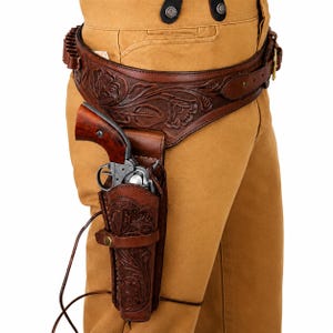 May include: A brown leather gun holster and matching belt featuring intricate floral tooling. The holster contains a revolver with a wooden grip and a metallic frame. The belt includes a row of cartridge loops and is worn over tan trousers.