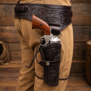 Könnte beinhalten: Dunkelbraunes Leder-Gürtel- und Holster-Set mit floralen Verzierungen. Das Holster hält einen silbernen Revolver mit Holzgriff. Der Gürtel hat Schlaufen für Kugeln. Das Set wird an der Taille und am Bein getragen.