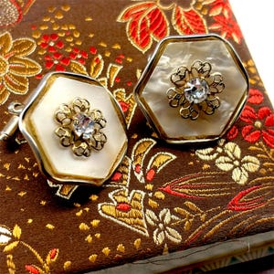 May include: A pair of vintage cufflinks with a hexagonal shape. Each cufflink features a cream-colored center with a gold-colored filigree flower and a clear crystal. The cufflinks have a silver-colored border and are set against a patterned background.