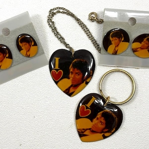 May include: A collection of Michael Jackson themed jewelry, including two pairs of earrings, a heart-shaped pendant necklace, and a heart-shaped keychain. The jewelry features a portrait of Michael Jackson.