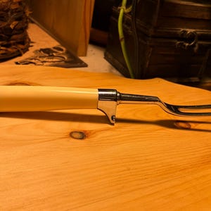 May include: A vintage carving fork with a cream-colored handle and a polished metal bolster and tines. The fork is designed for carving and serving meats. The handle has a smooth, rounded shape, and the tines are slender and sharp.