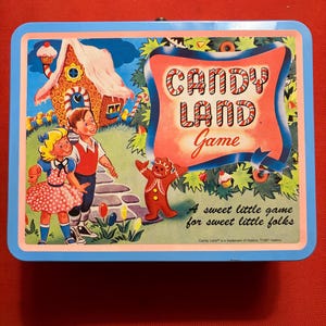 May include: A vintage Candy Land game tin with a blue border. The lid features a colorful illustration of children, a gingerbread man, and a candy house. The text "CANDY LAND Game" is prominently displayed.