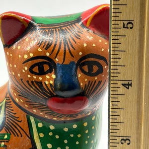 May include: A hand-painted ceramic cat figurine. The cat has a brown body with black and yellow details, red ears, a blue nose, and a red mouth. The cat's collar is green with white polka dots. The figurine is approximately 4 inches tall.
