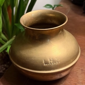 Puede incluir: Un jarrón de latón antiguo con un cuerpo redondeado y un borde ancho y acampanado. El jarrón tiene un tono dorado y una apariencia ligeramente envejecida. Las iniciales "L.H.P." están grabadas en el lateral. El jarrón probablemente se utiliza con fines decorativos.