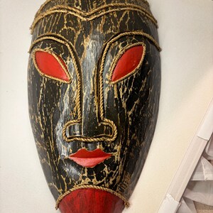 Op de afbeelding: Een decoratief wandmasker met een donkere, verweerde zwart-gouden afwerking, opvallende rode amandelvormige ogen, rode lippen en een rode kin. De gelaatstrekken zijn omlijnd met dikke, natuurkleurige gedraaide touwdetails.