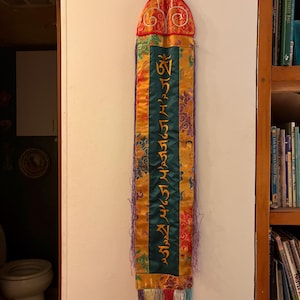 May include: A decorative wall hanging with a red and gold top, featuring ornate designs. The main body is a mix of gold, green, and patterned fabrics, with gold lettering. The bottom has colorful fringe.