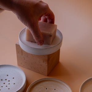 Puede incluir: Tres jaboneras de cerámica con agujeros de drenaje. Dos son blancas con un esmalte marrón claro y una es marrón claro con un esmalte blanco. Una pastilla de jabón está sobre una de las jaboneras.