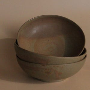 Puede incluir: Tres cuencos de cerámica apilados en tonos marrones y verdes. Los cuencos están esmaltados y tienen un aspecto rústico y hecho a mano.