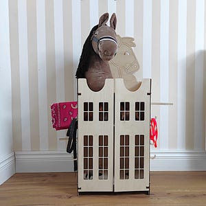 May include: A wooden playhouse for children with a brown plush horse inside. The playhouse has a red bandana and a black leather strap hanging on the side. The playhouse has two windows on each side and a small roof.
