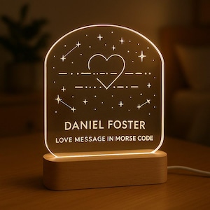 Może przedstawiać: Spersonalizowana lampka nocna z drewnianą podstawą i przezroczystym panelem akrylowym. Panel przedstawia serce, gwiazdy i imię "DANIEL FOSTER" z tekstem "LOVE MESSAGE IN MORSE CODE". Światło emituje ciepły blask.