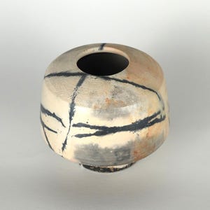 Puede incluir: Un jarrón de cerámica con cuerpo redondo y una pequeña abertura en la parte superior. El jarrón es de color blanquecino con marcas abstractas negras y grises. La base tiene un aspecto texturizado marrón oscuro. Adecuado para la decoración del hogar.