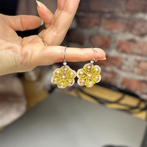 Könnte beinhalten: Ein Paar blütenförmige Ohrringe mit beigefarbenen und goldenen Perlen. Die Ohrringe sind mit einem gehäkelten Design gefertigt und haben silberne Haken.