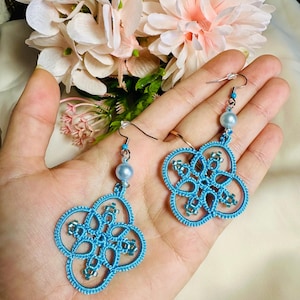 Könnte beinhalten: Ein Paar blaue gehäkelte Ohrringe mit einer Perle an jedem Ohrring. Die Ohrringe haben die Form eines vierzackigen Sterns.