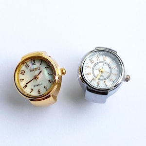 Può includere: Due orologi ad anello. Uno è dorato con quadrante bianco e le parole "ROZHI" e "QUARTZ" stampate. L'altro è argentato con quadrante bianco e numeri neri.