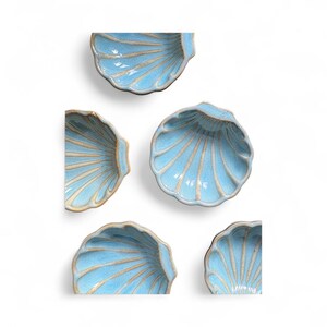 May include: A collection of five light blue ceramic shell-shaped dishes. Each dish has a textured surface with radial lines and a contrasting tan rim. The dishes are arranged on a white background.
