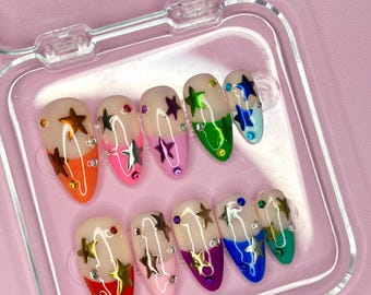 Puede incluir: Un juego de diez uñas postizas con un diseño degradado arcoíris y detalles de estrellas doradas. Las uñas se muestran en un estuche de plástico transparente.