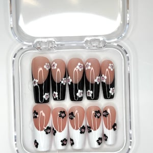 Può includere: Un set di unghie finte in una custodia di plastica trasparente. Le unghie presentano un design con base nude, punte nere e accenti floreali bianchi e neri. I fiori hanno piccole gemme rosa al centro.