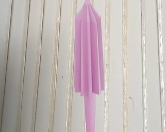 May include: A light purple candle with a unique, draped design. The candle has a flared top and a long, cylindrical base. The background is a white, vertical-striped surface.