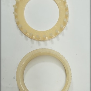 May include: Two beige plastic knitting looms are shown against a white backdrop. The top loom is a ring with pegs and numbers. The bottom loom is a ring with a ridged edge. These tools are used for knitting.