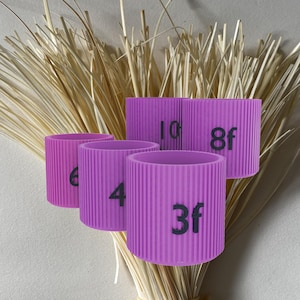 Peut inclure: Quatre anneaux en silicone violet avec des chiffres noirs imprimés dessus. Les chiffres sont 6, 4, 3f et 8f. Les anneaux sont posés sur un tas de paille.