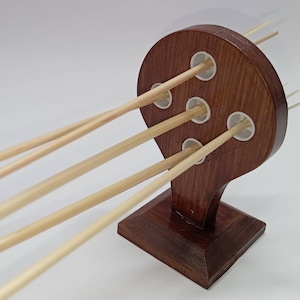 May include: A wooden incense holder with a dark brown finish. The holder features a rounded top with five holes, each containing a white insert. Several thin, light-colored incense sticks are inserted into the holes, angled outwards.