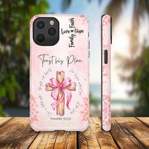 May include: A pink phone case with a cross design and the text "Trust his Plan." The cross is adorned with a pink bow. Additional text includes "Love, Hope, Family, Faith" and "Proverbs 31:25."