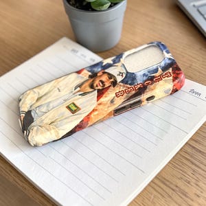 May include: A phone case with a portrait of a person in a white jacket against a red and blue backdrop. The case sits on a lined notepad on a wooden desk, with a small potted succulent in the background.