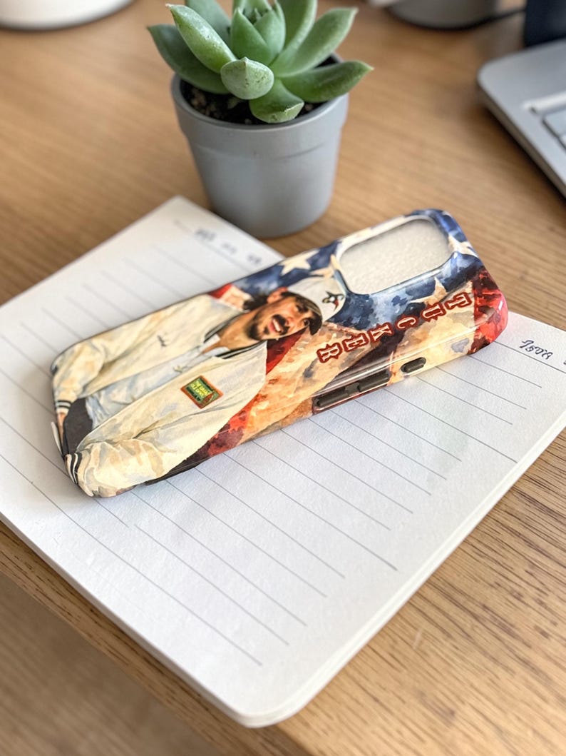 May include: A phone case with a portrait of a person in a white jacket against a red and blue backdrop. The case sits on a lined notepad on a wooden desk, with a small potted succulent in the background.