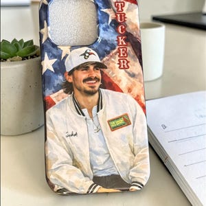 May include: A phone case with a watercolour illustration of a person in a white jacket and baseball cap. The background features a painted American flag design with the name "TUCKER" vertically. The jacket has the word "Jacket" on it.