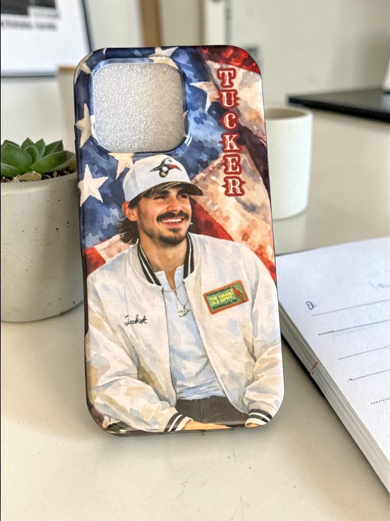 May include: A phone case with a watercolour illustration of a person in a white jacket and baseball cap. The background features a painted American flag design with the name "TUCKER" vertically. The jacket has the word "Jacket" on it.