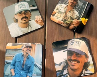 May include: Four square coasters with images of a man wearing hats and casual shirts. The coasters are on a wooden surface. The man is wearing a white t-shirt, a denim shirt, and a patterned shirt. The coasters have different hat designs.