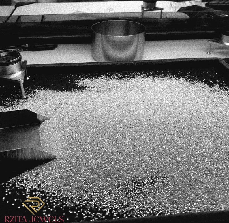 Pode incluir: Imagem a preto e branco de uma bandeja cheia de pequenas pedras preciosas brilhantes. Uma tigela de metal e instrumentos de medi&ccedil;&atilde;o s&atilde;o vis&iacute;veis no fundo. O logotipo "RZITA JEWELS" est&aacute; no canto inferior esquerdo.