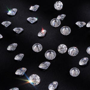 Pode incluir: Uma cole&ccedil;&atilde;o de diamantes brilhantes e transparentes espalhados sobre uma superf&iacute;cie preta. Os diamantes variam em forma, com pedras redondas e em forma de p&ecirc;ra. A imagem &eacute; bem iluminada, destacando as facetas e o brilho das pedras preciosas. O logotipo RZITA JEWELS &eacute; vis&iacute;vel no canto inferior esquerdo.