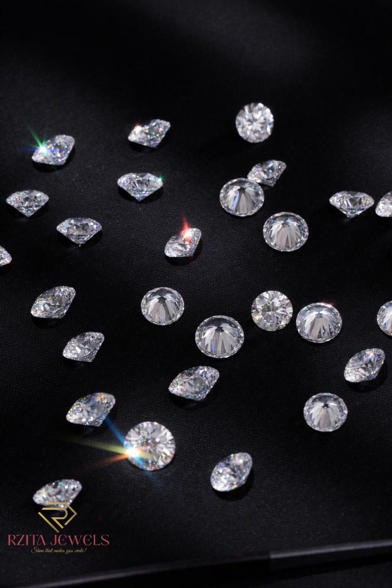 Pode incluir: Uma cole&ccedil;&atilde;o de diamantes brilhantes e transparentes espalhados sobre uma superf&iacute;cie preta. Os diamantes variam em forma, com pedras redondas e em forma de p&ecirc;ra. A imagem &eacute; bem iluminada, destacando as facetas e o brilho das pedras preciosas. O logotipo RZITA JEWELS &eacute; vis&iacute;vel no canto inferior esquerdo.