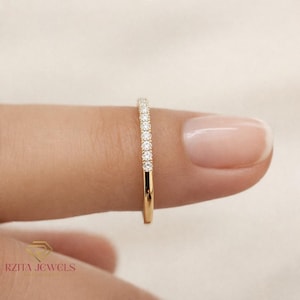 May include: A gold band ring with a row of small, clear gemstones set along the top. The ring is held between two fingers, showcasing the delicate design and sparkle of the stones. The ring is a classic piece of jewelry.