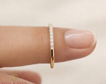 Pode incluir: Um anel de banda de ouro com uma fileira de pequenas pedras preciosas claras colocadas na parte superior. O anel é segurado entre dois dedos, mostrando o design delicado e o brilho das pedras. É uma joia clássica.