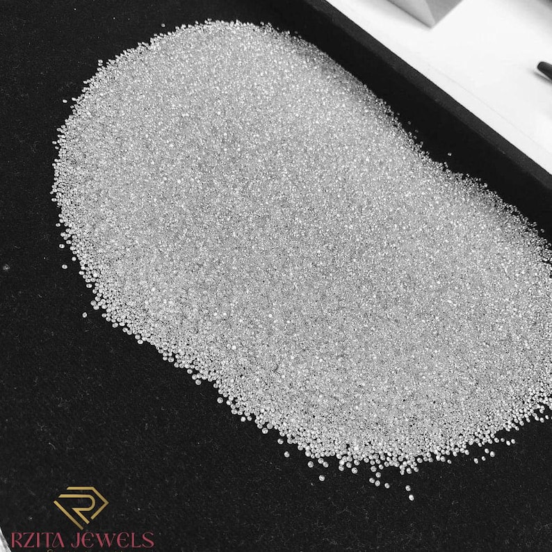 Pode incluir: Uma grande pilha de pequenas pedras preciosas redondas, claras e brilhantes, possivelmente diamantes, preenche uma bandeja preta. As pedras preciosas s&atilde;o densamente embaladas, refletindo a luz e criando uma exibi&ccedil;&atilde;o brilhante. O logotipo "RZITA JEWELS" &eacute; vis&iacute;vel no canto inferior esquerdo.