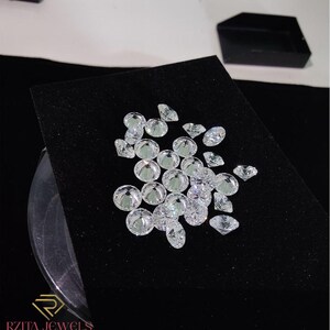 Pode incluir: Uma cole&ccedil;&atilde;o de diamantes claros e brilhantes de v&aacute;rias formas e tamanhos, incluindo cortes redondos e ovais, &eacute; exibida em uma superf&iacute;cie de veludo preto. Os diamantes refletem a luz, criando uma exibi&ccedil;&atilde;o brilhante. Uma caixa de joias preta est&aacute; no fundo.