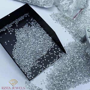 Pode incluir: Uma foto em close-up de in&uacute;meros diamantes pequenos e transparentes espalhados sobre uma superf&iacute;cie branca e dentro de uma bandeja preta. Os diamantes brilham, refletindo a luz. Uma sacola pl&aacute;stica cheia de mais diamantes &eacute; vis&iacute;vel ao fundo. O logotipo "RZITA JEWELS" est&aacute; no canto inferior esquerdo.