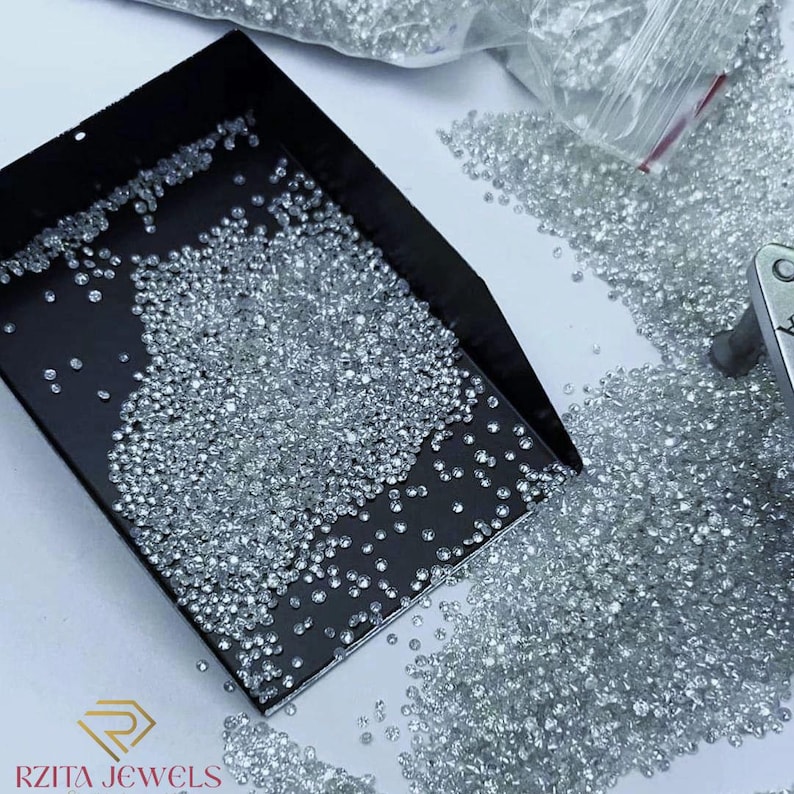 Pode incluir: Uma foto em close-up de in&uacute;meros diamantes pequenos e transparentes espalhados sobre uma superf&iacute;cie branca e dentro de uma bandeja preta. Os diamantes brilham, refletindo a luz. Uma sacola pl&aacute;stica cheia de mais diamantes &eacute; vis&iacute;vel ao fundo. O logotipo "RZITA JEWELS" est&aacute; no canto inferior esquerdo.