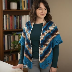 May include: A blue, brown, and white crocheted shawl with a scalloped edge. The shawl is worn over a dark teal long-sleeved shirt and gray pants. The background includes a bookshelf and a white couch.