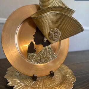 May include: A decorative gold charger plate featuring a black silhouette of a woman wearing a gold hat with a floral embellishment. The woman's hands are clasped in prayer. The plate is displayed on a gold decorative plate.